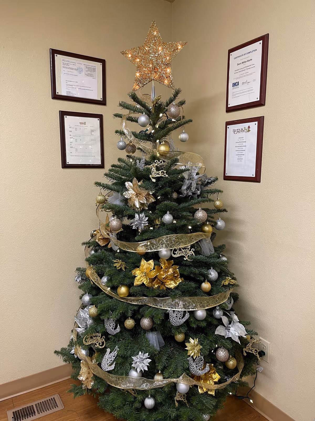 A decorated Christmas tree placed in the corner of a room with beige walls. The tree is adorned with gold, silver, and white ornaments, including baubles, flowers, ribbons, and a lit star on top. Four framed certificates or documents are hung on the walls behind the tree.