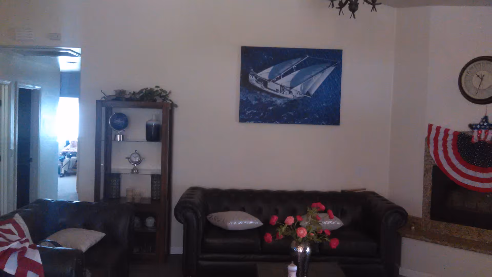 A living room with two dark leather sofas, one with two light-colored pillows and a vase with pink flowers on a small table in front. There is a wooden shelf with decorative items and a plant on top, a painting of a sailboat on the wall, and a fireplace decorated with a patriotic red, white, and blue banner and a clock above it.
