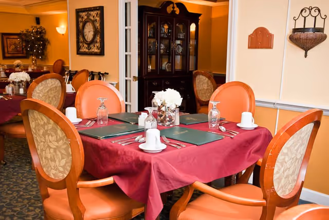 A dining room table set for four with a burgundy tablecloth, green placemats, white cups and saucers, upside-down water glasses, silverware, and a centerpiece of white flowers. The room has warm yellow walls, upholstered wooden chairs, a clock on the wall, and a dark wooden cabinet in the background.