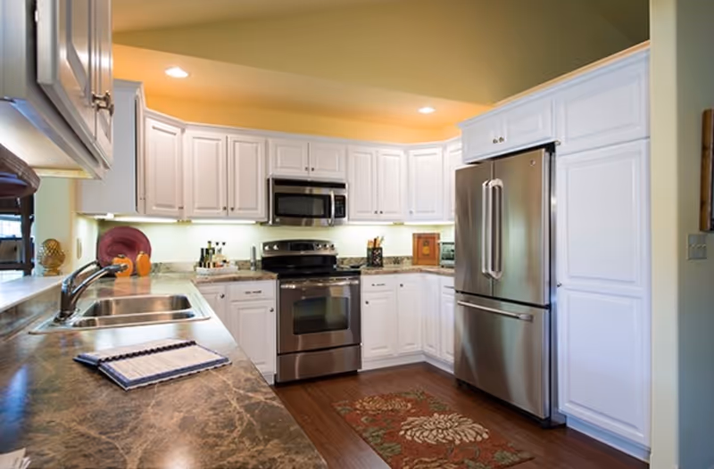 A modern kitchen with white cabinets, stainless steel appliances including a refrigerator, oven, and microwave. The kitchen features a marble countertop with a sink and a decorative rug on the wooden floor. The walls are painted light green and the ceiling has recessed lighting.