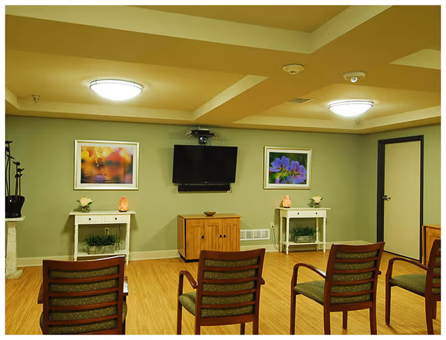 A small common area with four wooden chairs facing a wall-mounted flat screen TV. The walls are painted light green and decorated with two framed floral pictures. Below the TV are two small white tables with plants and decorative items, and a wooden cabinet. The ceiling has recessed lighting fixtures.