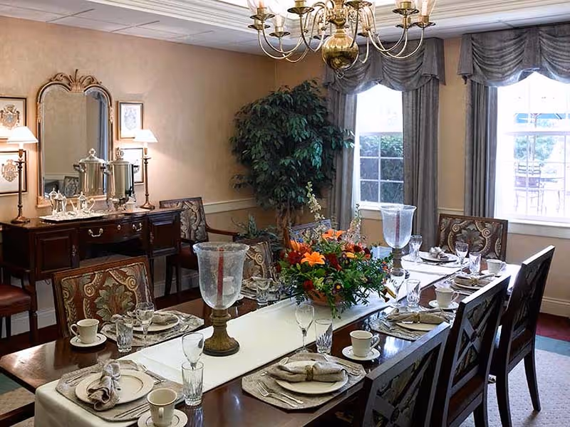 A formal dining room with a long wooden table set for eight people. The table is decorated with a white runner, a large floral centerpiece, glassware, plates with napkins, and cups. There are upholstered chairs around the table. In the background, there is a sideboard with silver tea sets and two lamps, a large mirror, a potted plant, and windows with draped curtains letting in natural light. A chandelier hangs above the table.