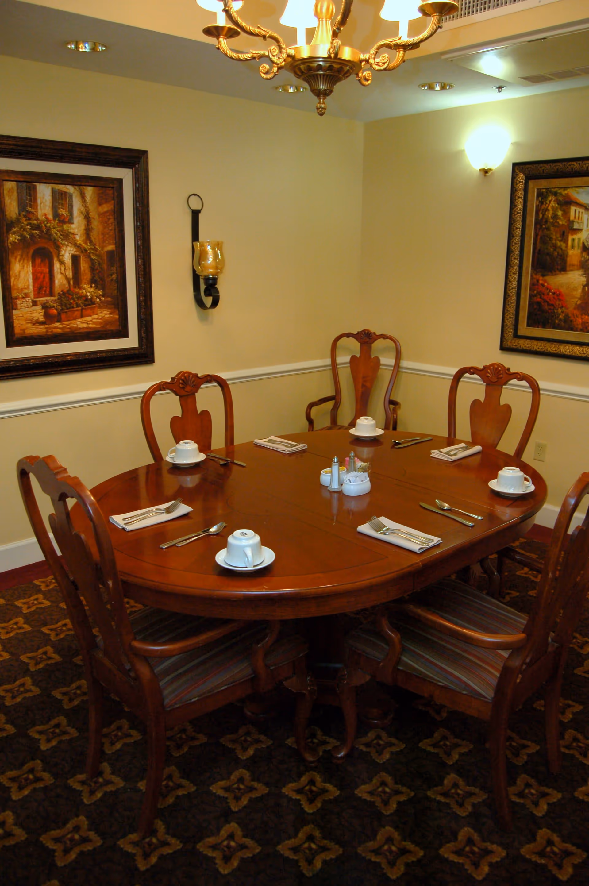 A formal dining room with a polished wooden oval table set for six people. Each place setting includes a white cup and saucer, a folded napkin with a fork and knife, and a spoon. The room has beige walls adorned with framed paintings and a decorative wall sconce. A chandelier hangs from the ceiling, and the floor is covered with a patterned carpet.