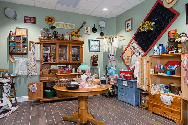 A cozy, vintage-style interior room with a round wooden table and cabinets filled with antiques and decorative decor against a green wall.