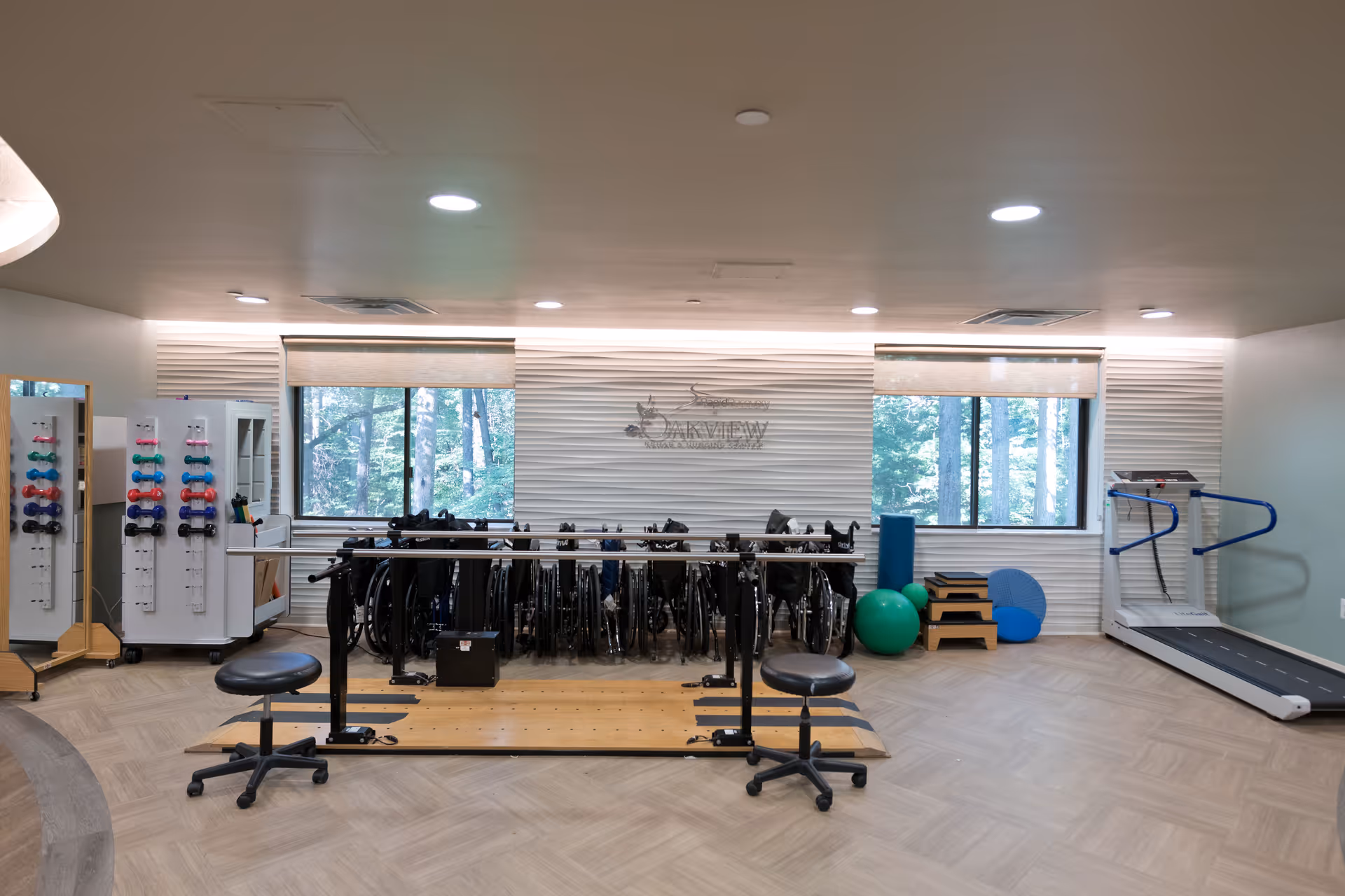 A rehabilitation or physical therapy room with parallel bars for walking exercises, two stools, a treadmill, exercise balls, and a rack of wheelchairs against a textured white wall with two windows showing trees outside.