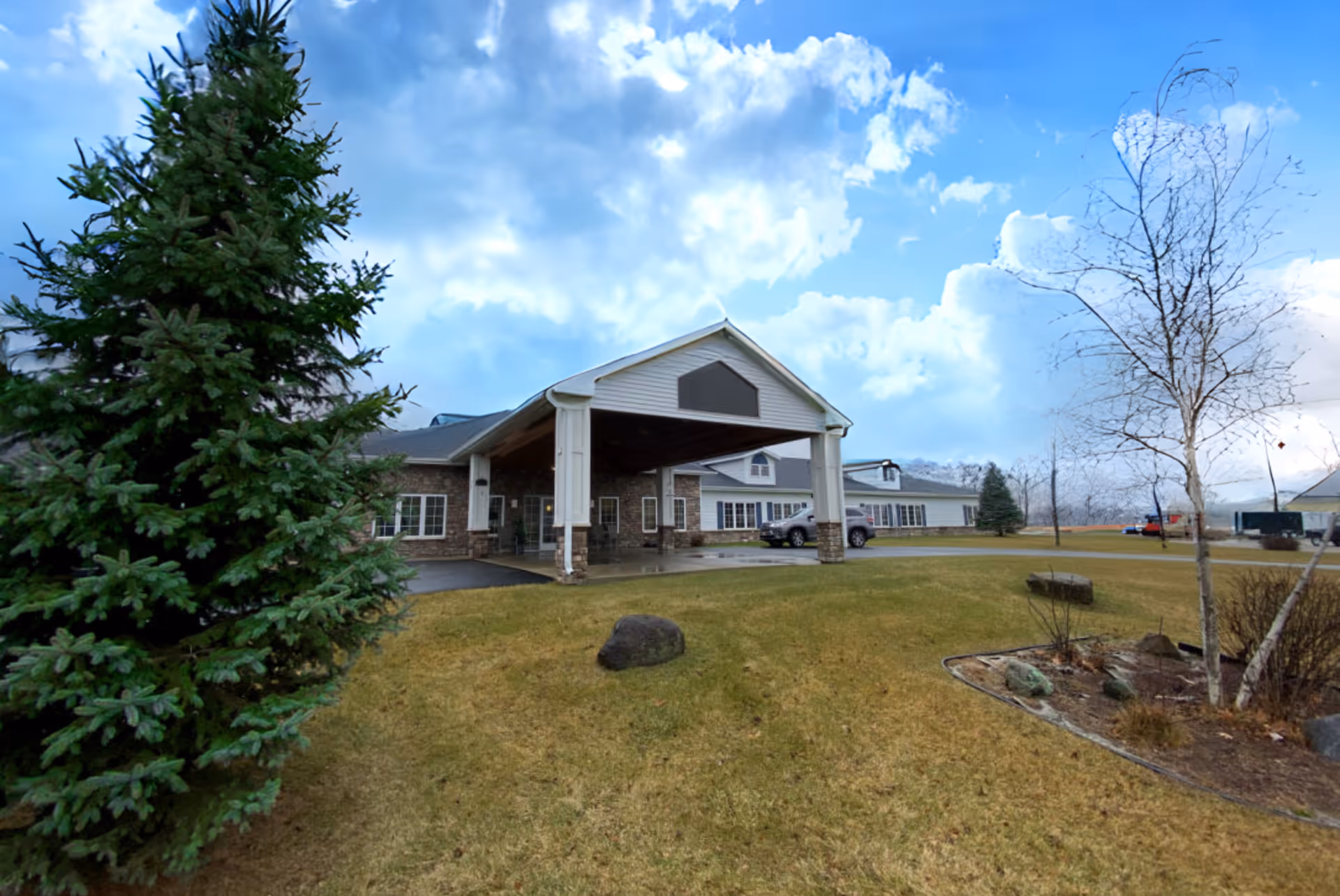 Exterior view of Charleston House Assisted Living facility showing the main entrance with a covered drop-off area, surrounded by a grassy lawn with trees and a partly cloudy sky.