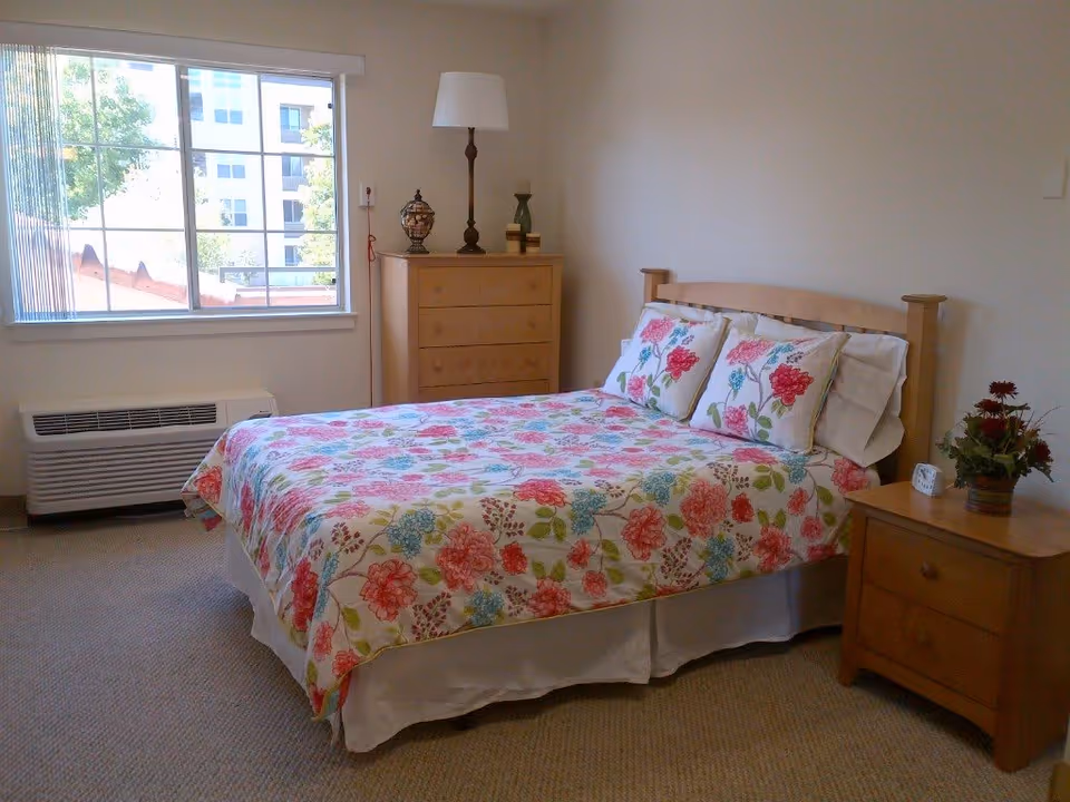 A bright bedroom with a double bed covered in floral bedding, wooden nightstand and dresser, and a large window.