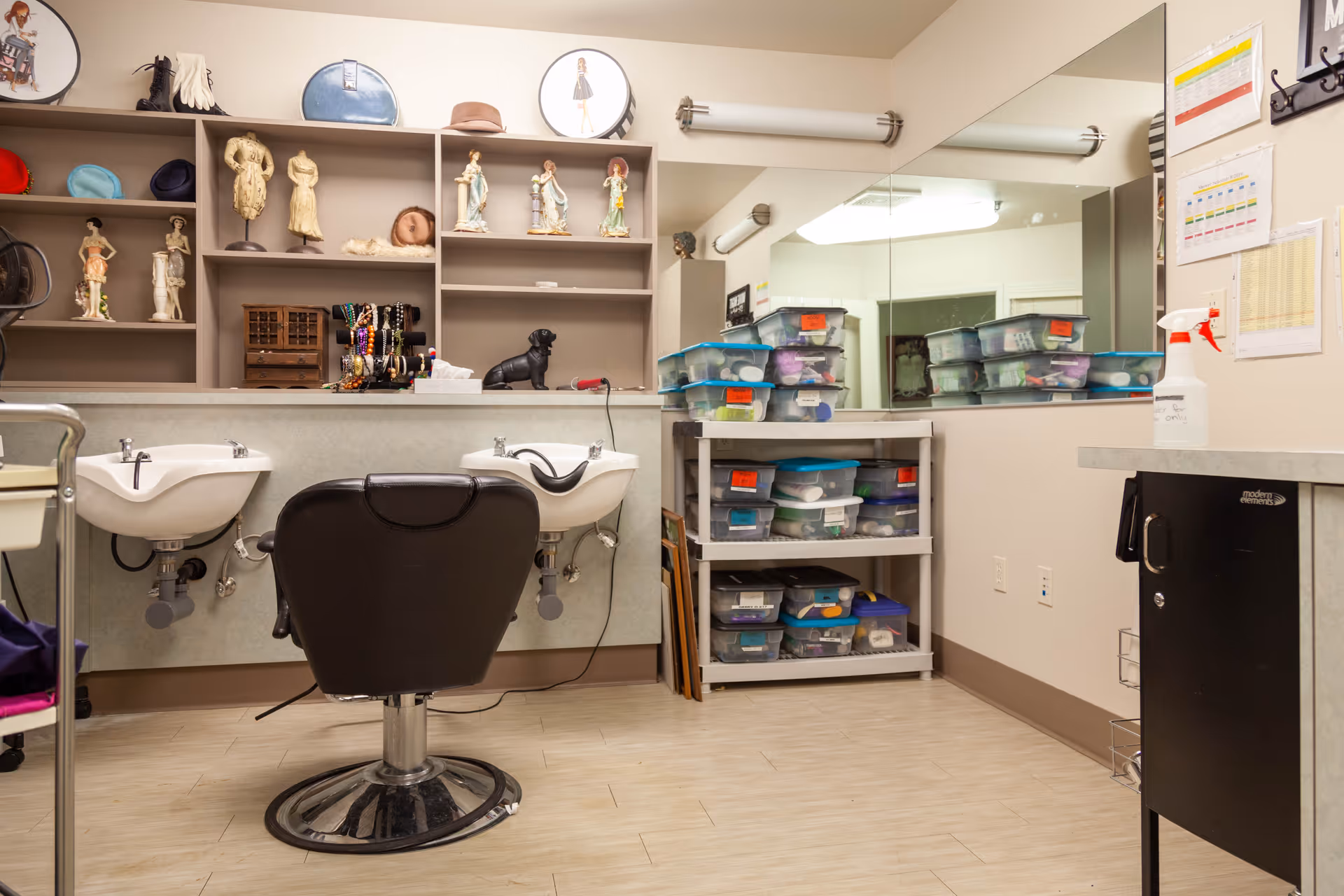 A small salon area with two white wash basins and a black salon chair in front. Behind the basins are shelves displaying various decorative figurines, hats, and accessories. To the right, there is a shelving unit filled with clear plastic storage bins with blue lids. A large mirror covers the wall above the shelving unit and basins. The room has light-colored flooring and walls with some charts and a spray bottle on a counter to the right.