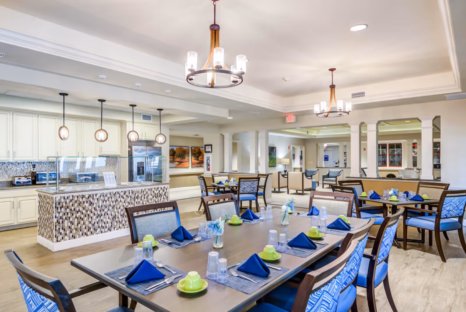 A bright and spacious dining room in Chantilly Heights Memory Care with multiple tables set with blue napkins, green cups and saucers, silverware, and clear plastic cups. The room features modern pendant lights, a serving counter with mosaic tile, cream-colored cabinetry, and comfortable seating with blue patterned cushions. The background shows a lounge area with chairs and artwork on the walls.