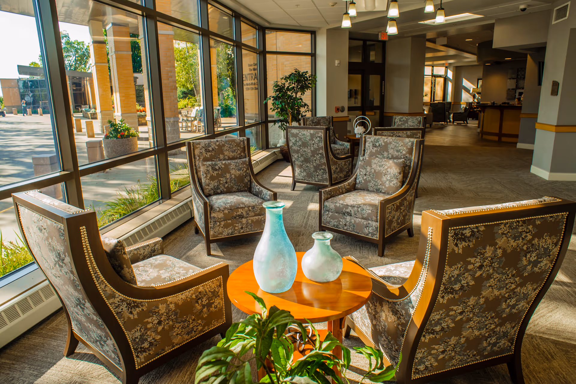 A bright and spacious seating area in a senior living facility with large windows letting in natural light. The area features several floral-patterned armchairs arranged around a small wooden table with two decorative glass vases. There are plants near the windows and the interior has a warm, inviting atmosphere with carpeted floors and overhead lighting.