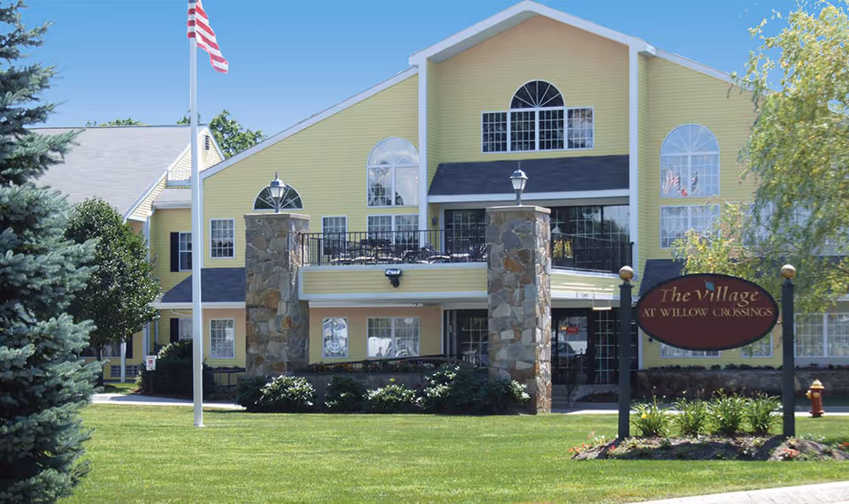 Exterior view of a large yellow building with stone pillars and multiple windows, a flagpole with an American flag, and a sign that reads 'The Village at Willow Crossings' on a well-maintained lawn with trees and shrubs.