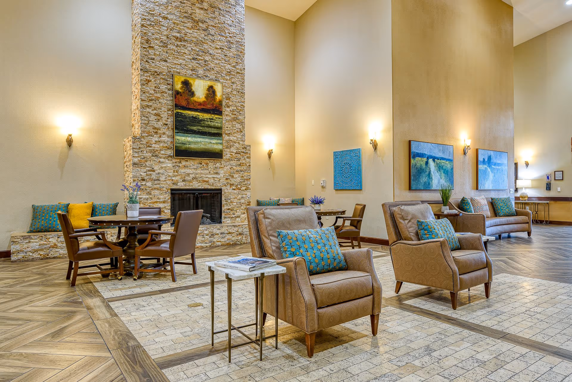 A spacious senior living facility common area with comfortable armchairs and sofas adorned with patterned cushions. The room features a tall stone fireplace with a colorful painting above it, round wooden tables with chairs, and decorative wall art. Warm lighting from wall sconces creates a cozy atmosphere.