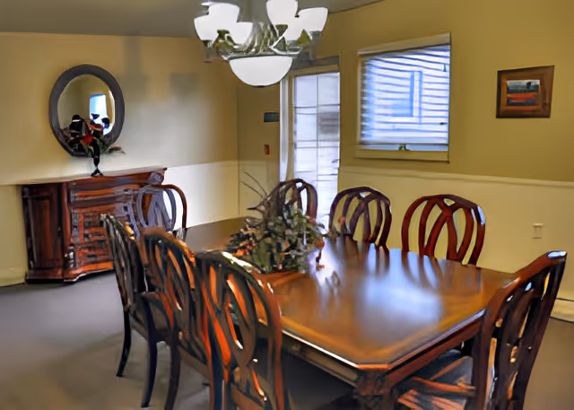 A dining room with a large wooden dining table surrounded by eight matching wooden chairs. A decorative centerpiece with greenery is placed on the table. On the left side of the room, there is a wooden sideboard with a vase of flowers and a round mirror hanging above it. The room has beige walls with a white chair rail, a window with blinds, a door with a glass panel, and a ceiling light fixture.