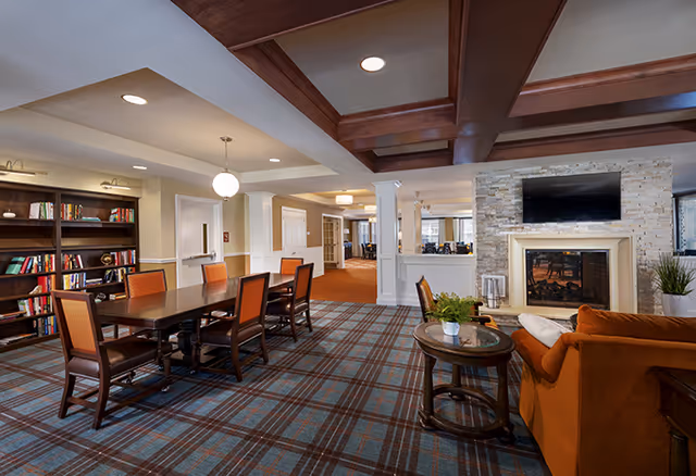 A spacious senior living facility common area featuring a long wooden table with six chairs, a bookshelf filled with books, a stone fireplace with a mounted flat-screen TV above it, and a comfortable orange sofa next to a round wooden coffee table with a small plant. The room has a patterned carpet, white walls with wooden ceiling beams, and soft lighting fixtures.
