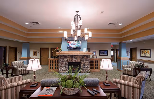 A cozy living room area in a senior living facility featuring a stone fireplace with a TV mounted above it. The room has multiple striped armchairs and side tables with lamps. A coffee table in the foreground holds a basket with green plants, a magazine, and remote controls. The ceiling has recessed lighting and a chandelier.