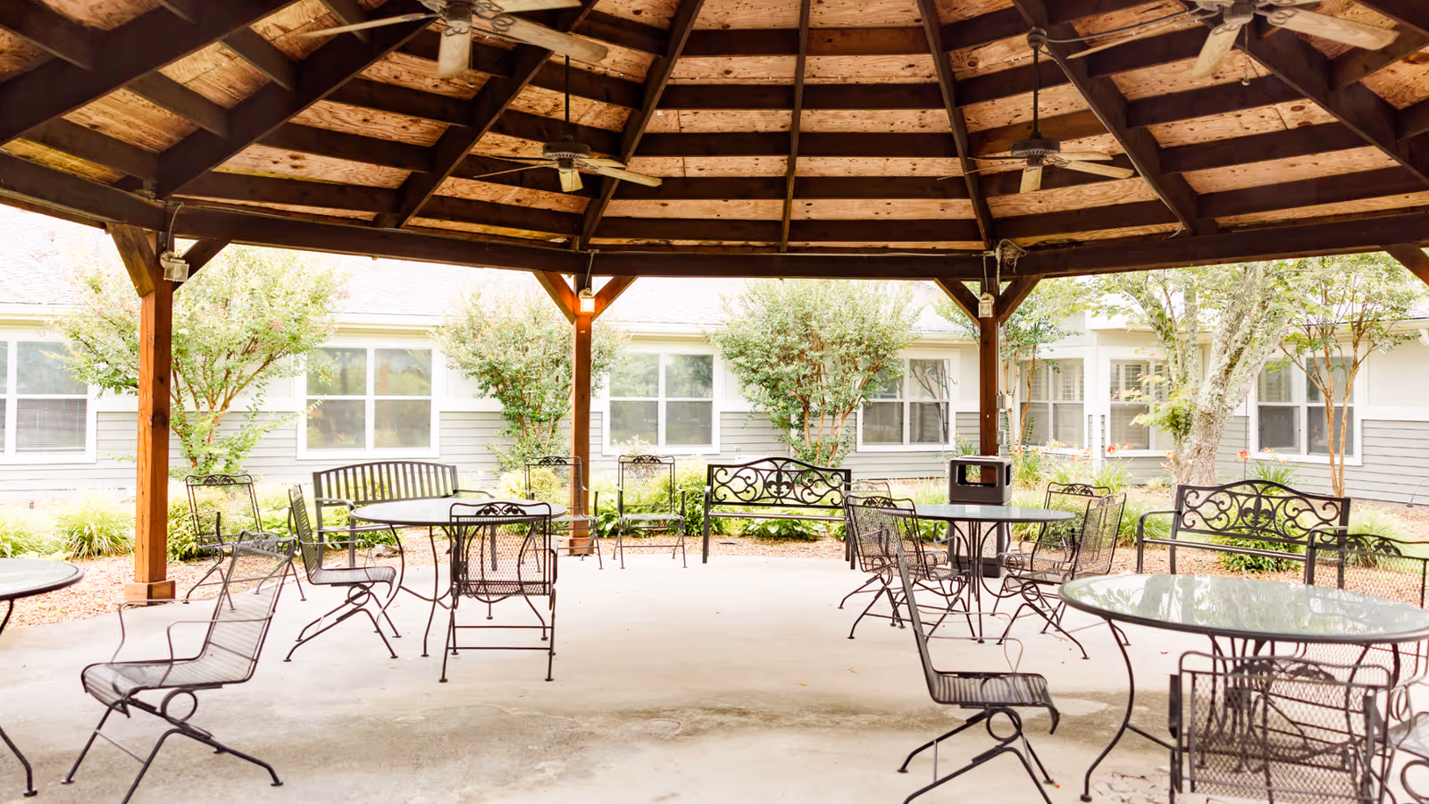 Outdoor covered seating area with metal tables and chairs under a wooden gazebo roof, surrounded by greenery and windows of a building in the background.