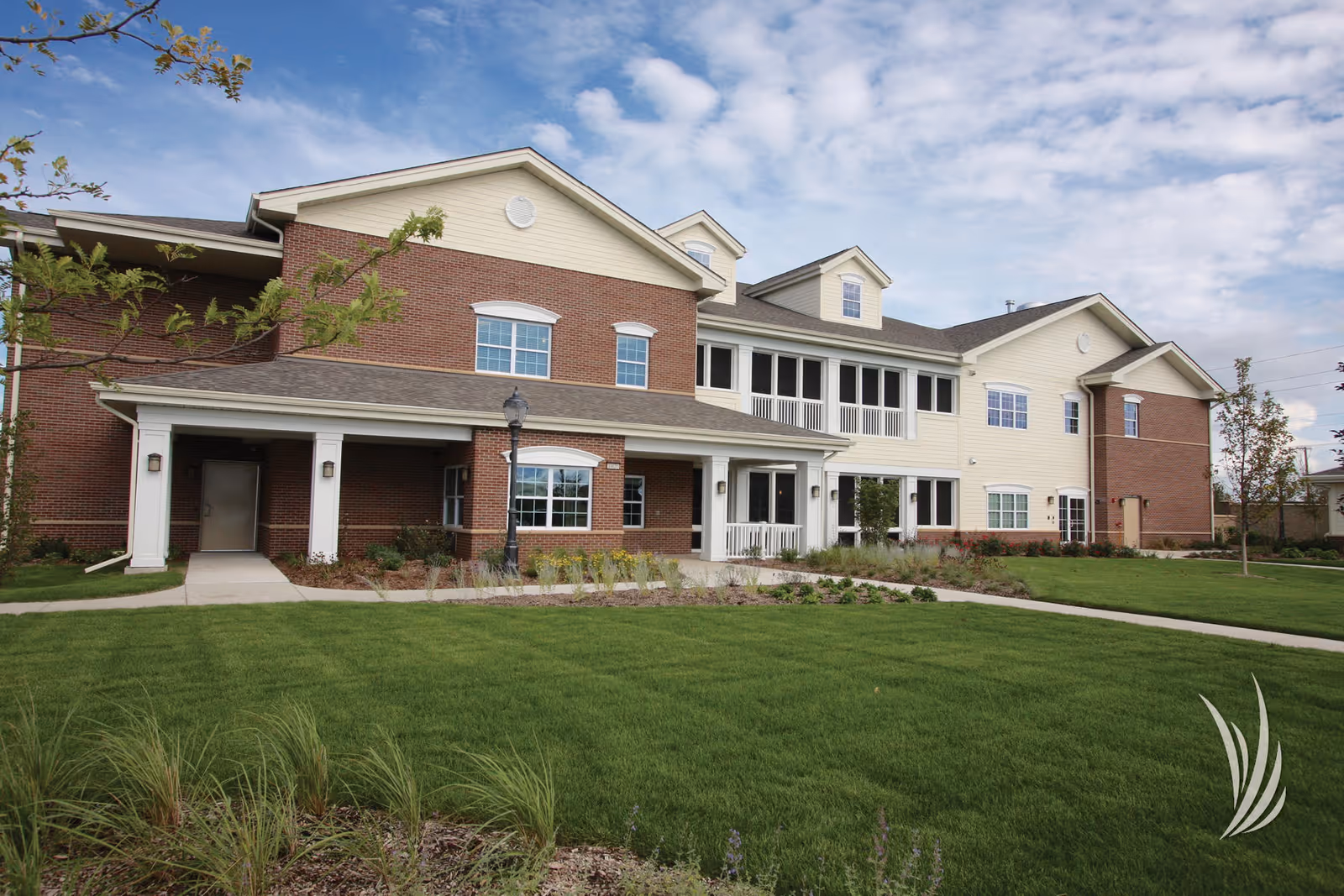 Two-story red-brick and light-sided senior living building with a covered entrance, porch, and manicured lawn under a partly cloudy sky.