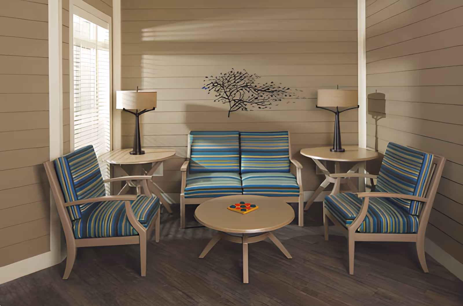 A cozy seating area with two armchairs and a loveseat, all featuring blue and green striped cushions. There are two round wooden side tables with matching lamps on either side of the loveseat, and a round wooden coffee table in the center with a small colorful game on it. The walls are beige with horizontal paneling, and a decorative metal wall art piece resembling a tree branch is mounted above the loveseat. Light filters in through a window with blinds on the left side.