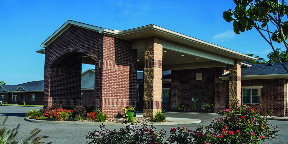Exterior view of Cabell Healthcare Center showing a brick building with a covered entrance supported by brick and stone pillars, surrounded by landscaped flower beds and a clear blue sky.