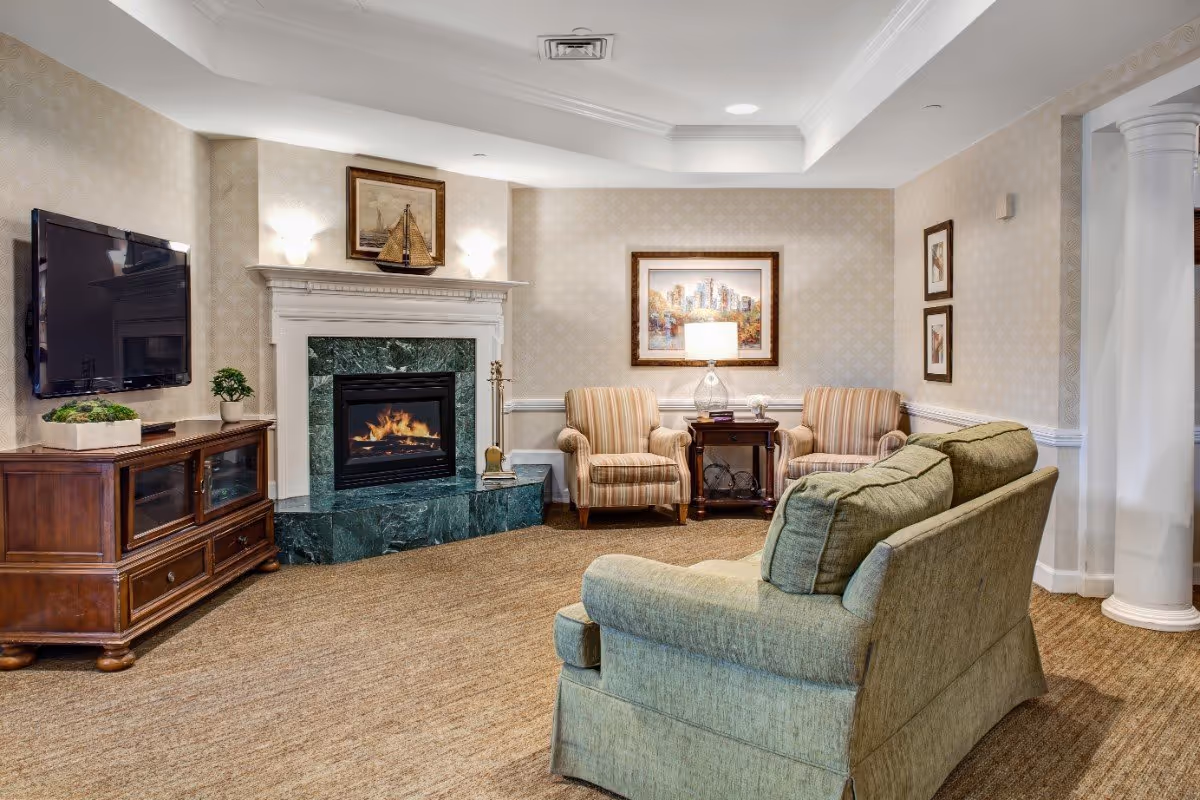 Cozy communal living room with a fireplace, wall-mounted TV, green sofa and striped armchairs around a side table.