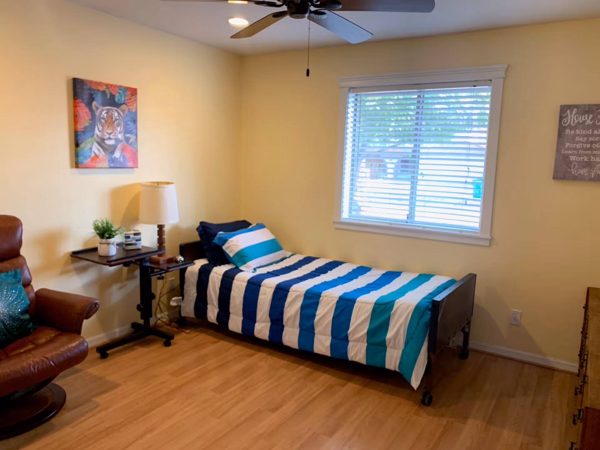 A simple bedroom with a single bed covered in a blue, white, and teal striped comforter. There is a brown leather recliner chair with a teal pillow, a small side table with a lamp and a plant, a ceiling fan, and a window with white blinds. The walls are painted light yellow and decorated with a colorful tiger painting and a framed inspirational quote.