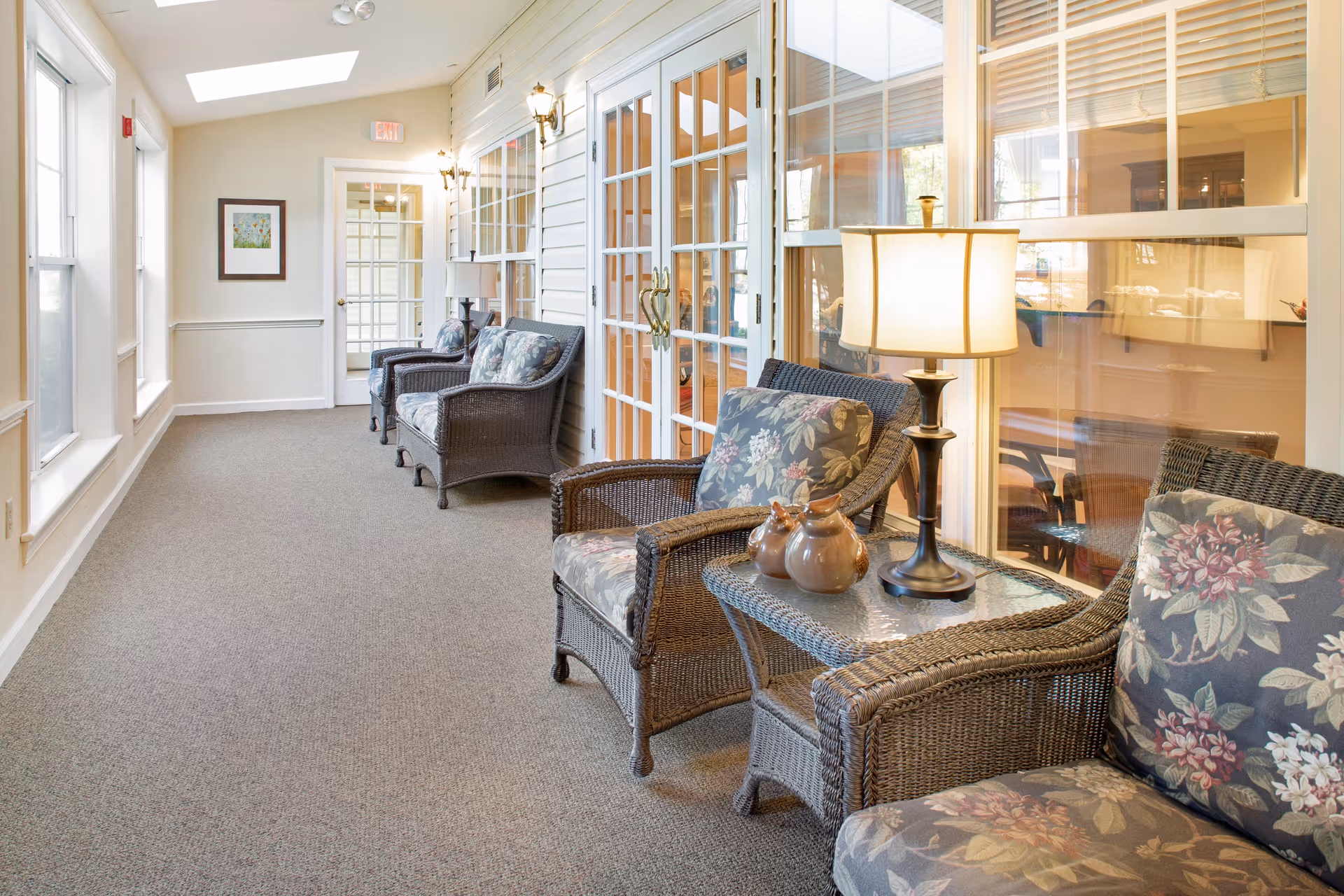 A bright indoor hallway with large windows on the left and a row of wicker chairs with floral cushions on the right. A glass-top side table with a lamp and decorative ceramic birds is placed between the chairs. The hallway has beige walls, carpeted floor, and glass-paneled doors leading to another room.