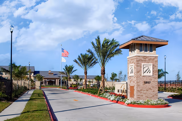 Entrance driveway to Mirador Retirement Community with a brick signpost displaying the community's logo and address number 5857. The driveway is lined with palm trees, flower beds, and street lamps under a partly cloudy blue sky. An American flag and another flag are flying on flagpoles near the entrance.