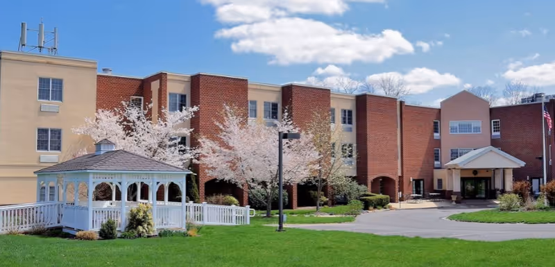 Exterior view of a senior living facility building with a combination of beige and red brick walls. In front of the building, there is a white gazebo surrounded by green grass and blooming trees under a blue sky with some clouds.