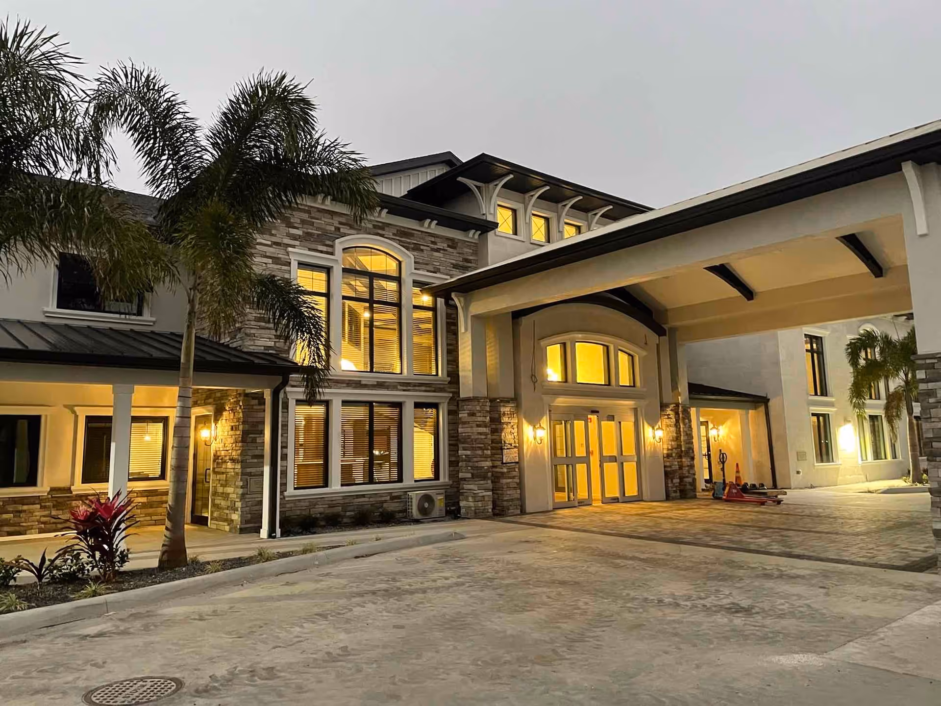 Exterior view of the Villas of Holly Brook building at dusk, showing a well-lit entrance with large windows, stone and stucco facade, palm trees, and a covered driveway.