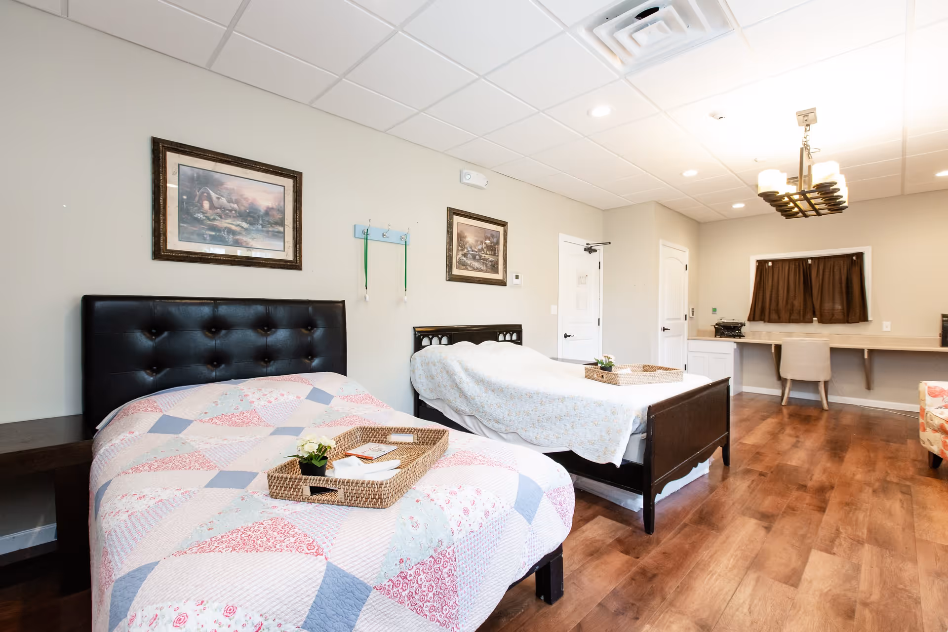 A bright, clean bedroom with two beds, each with a quilted bedspread and a wicker tray on top. The room has wooden flooring, two framed pictures on the wall, a desk with a chair, and a window with brown curtains.