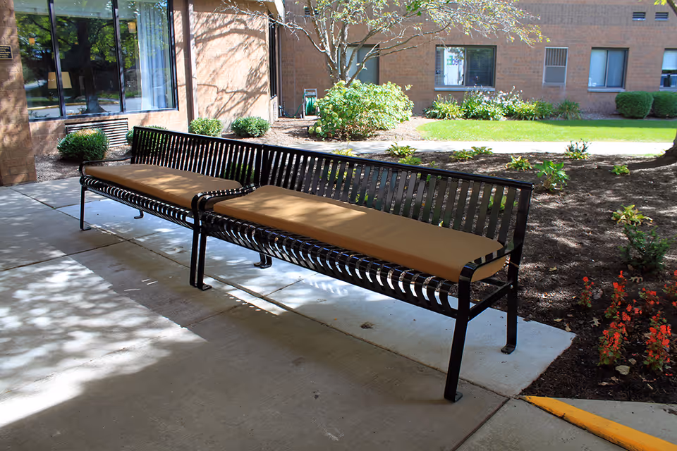 Two long black metal benches with tan cushions sit on a covered sidewalk outside a brick building amid landscaping.
