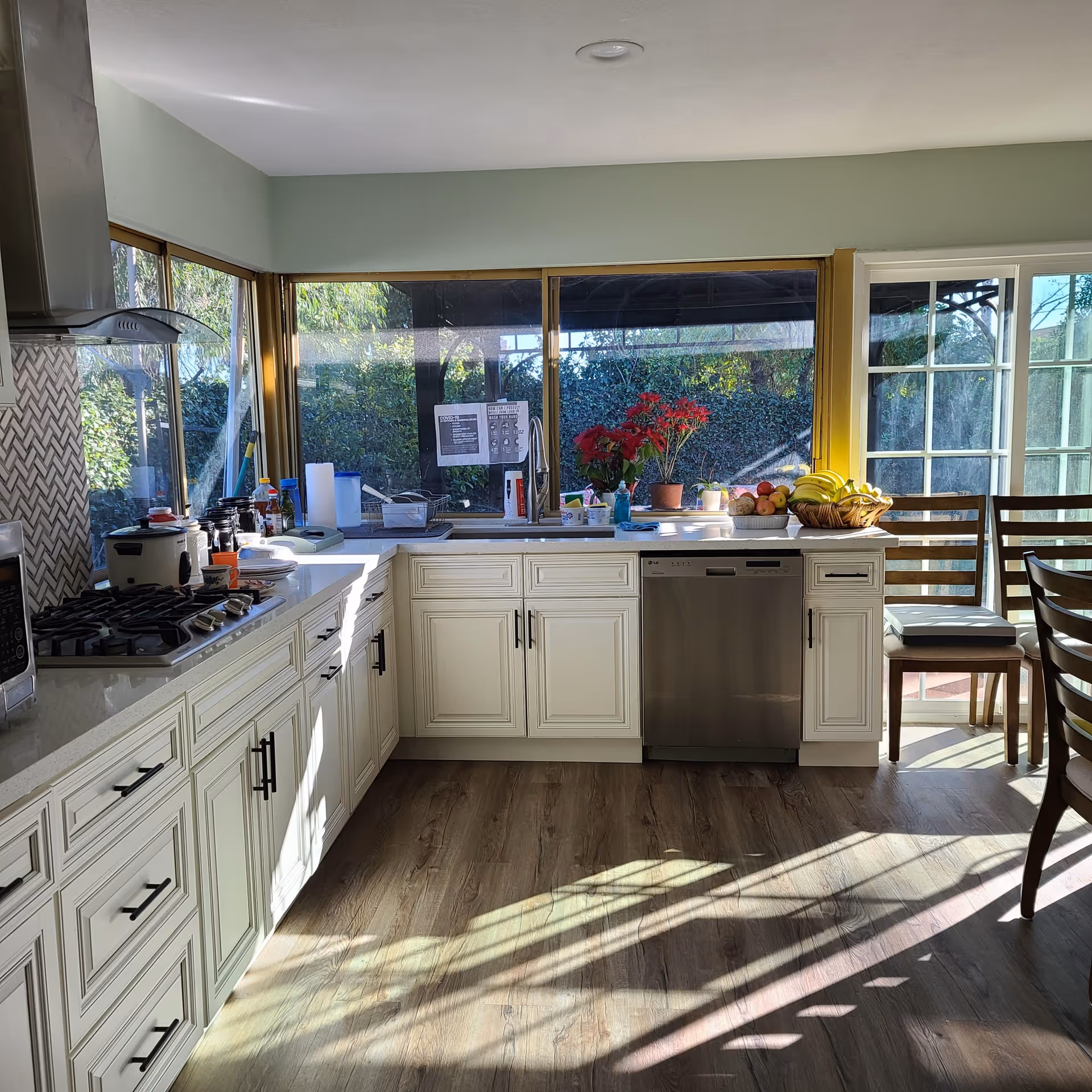 Bright kitchen with white cabinets, a gas stove, microwave, dishwasher, and large windows overlooking a garden. The countertop has various kitchen items including a rice cooker, fruit basket, and potted plants. Sunlight casts shadows on the wooden floor.