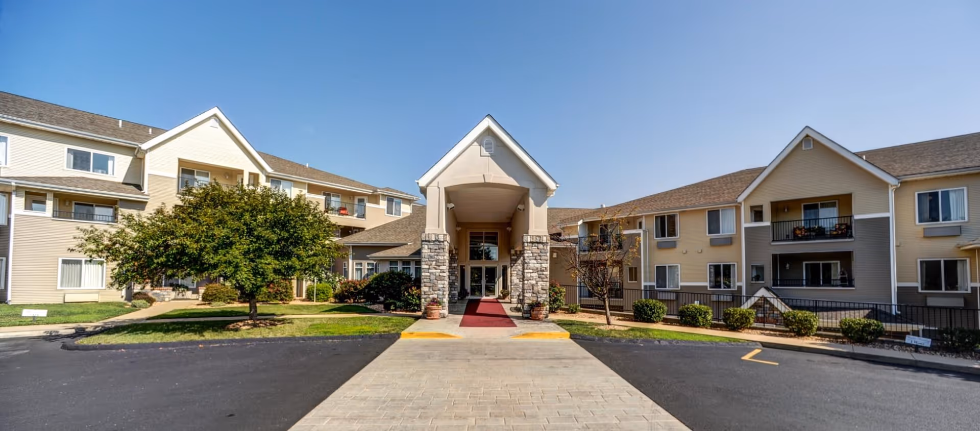 Front exterior view of College View Manor, a multi-story senior living facility with beige siding and stone accents around the entrance. The entrance has a covered portico with a red carpet leading to glass doors. There are trees, shrubs, and a paved driveway in front of the building under a clear blue sky.