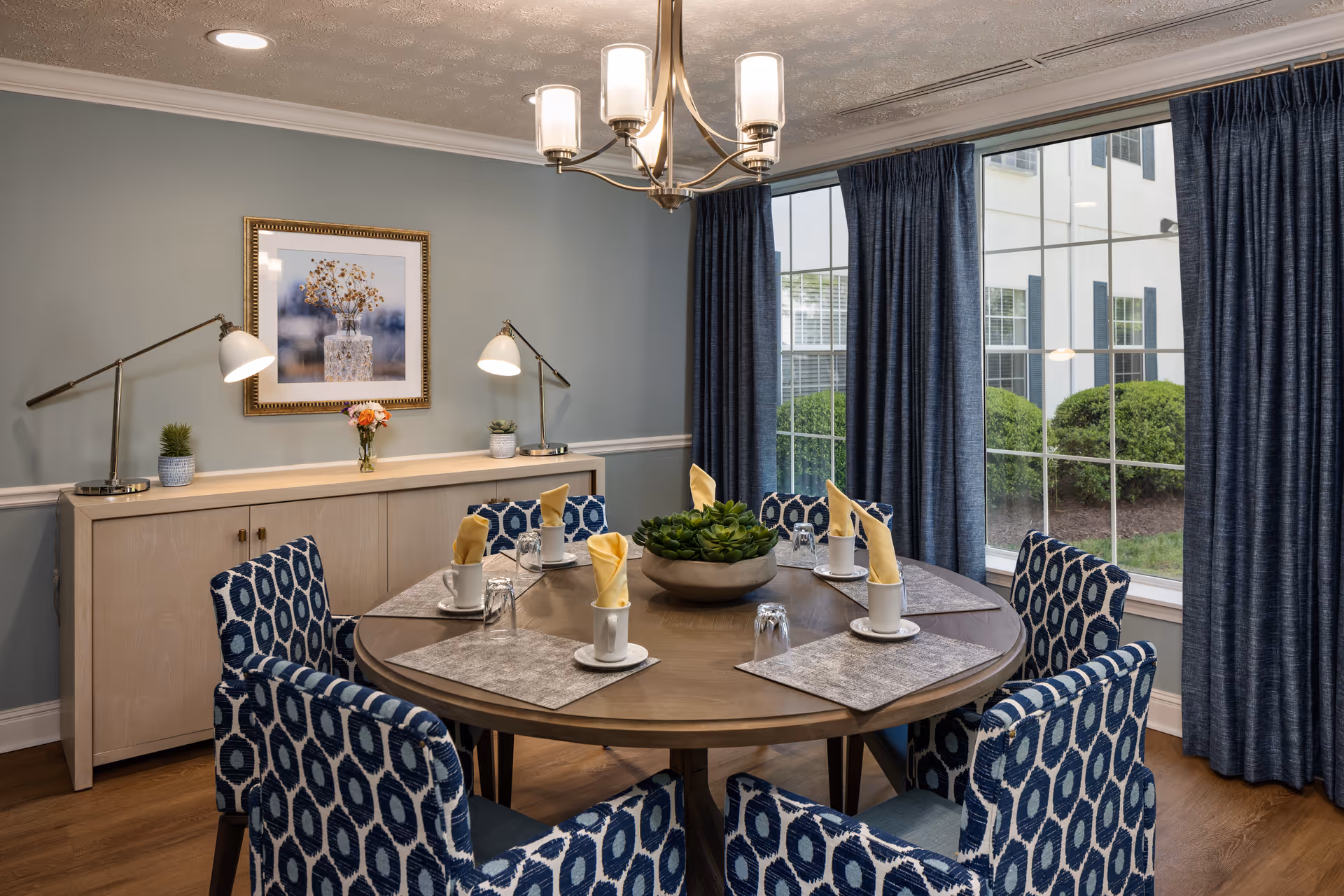 Well-appointed dining room with a round table set for six, patterned blue chairs, a buffet against a pale blue wall, and large windows with blue curtains.