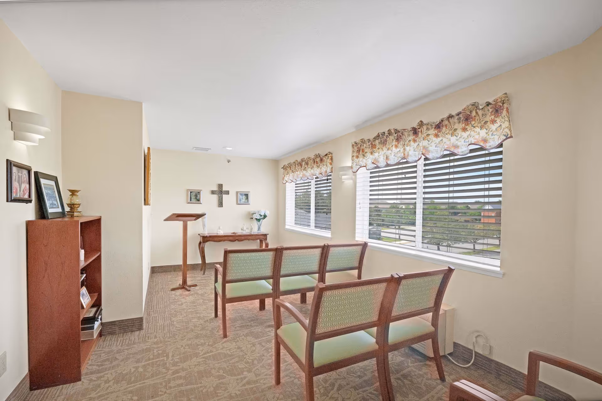 Small chapel-like room with rows of wooden chairs facing a lectern and a cross on the wall beside large windows with blinds and floral valances.