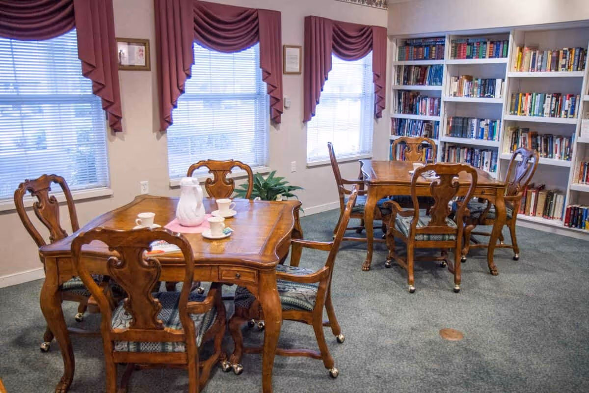 Wood-furnished common room with two wooden dining tables and chairs, bookshelves, and windows with mauve drapes.
