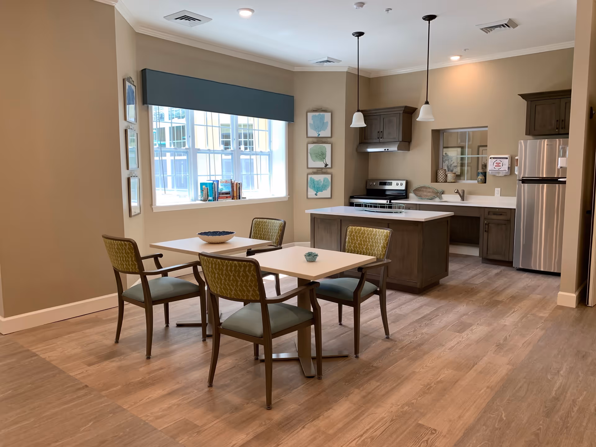 A bright and modern kitchen and dining area with light wood flooring, beige walls, and a large window with a blue valance. The kitchen features dark wood cabinets, a stainless steel refrigerator, an electric stove, and a white countertop island. In front of the window, there are two small square dining tables with four chairs that have green patterned backs and light blue seats. Three framed sea-themed artworks hang on the wall near the kitchen.