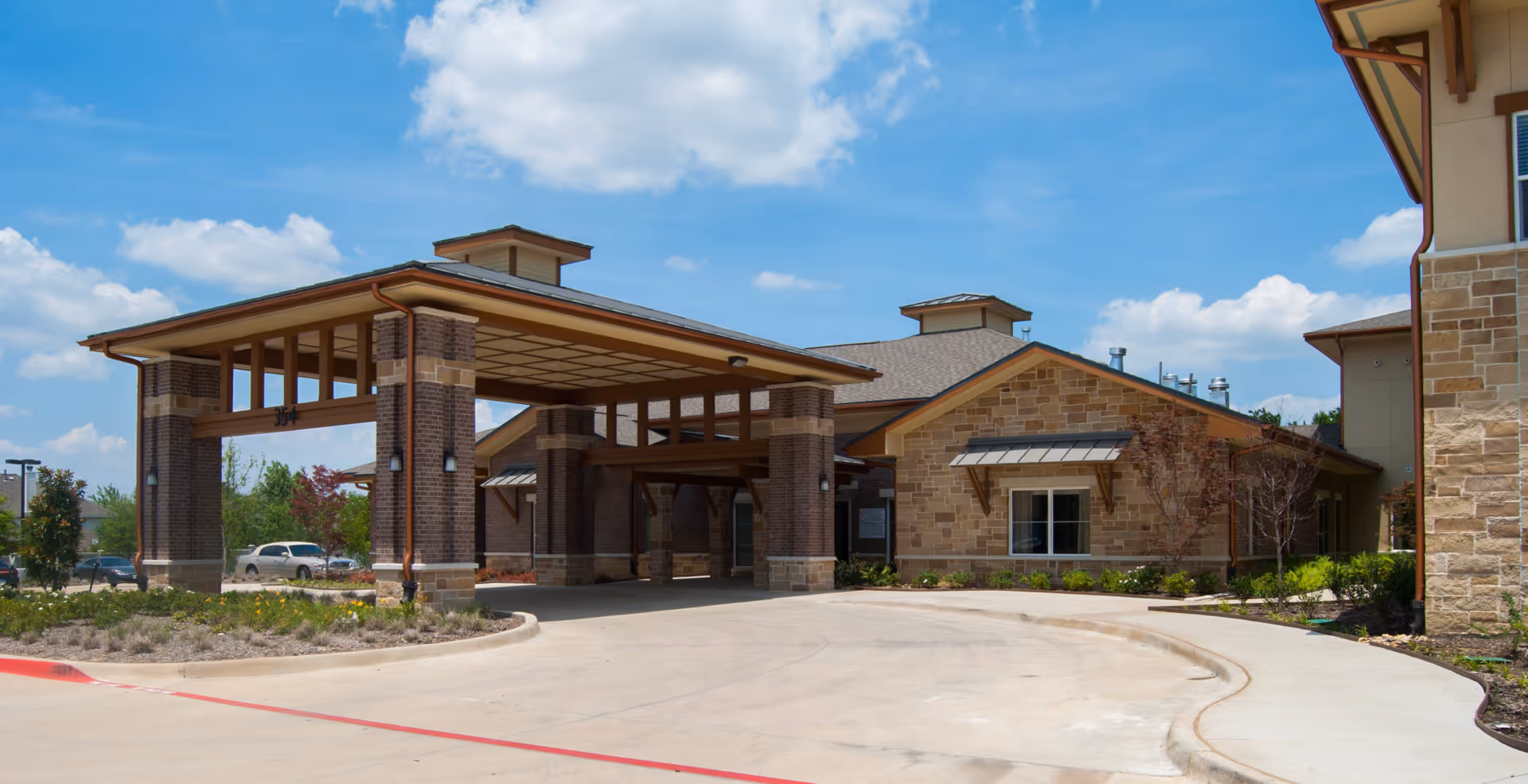 Exterior view of Cedar Bluff Assisted Living & Memory Care facility showing the main entrance with a covered drop-off area, brick and stone facade, and a clear blue sky with some clouds.
