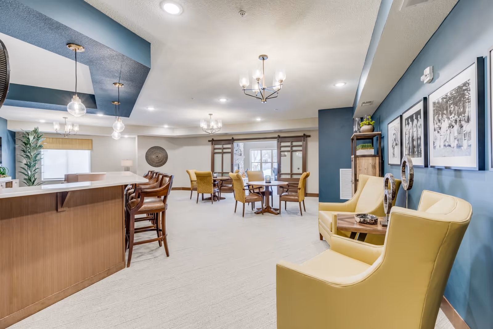 A spacious and well-lit common area in a senior living facility featuring a bar counter with wooden bar stools on the left, several round tables with yellow upholstered chairs in the center, and two yellow armchairs with a small wooden side table on the right. The walls are painted in blue and beige tones, decorated with black and white framed photographs and modern light fixtures hanging from the ceiling.