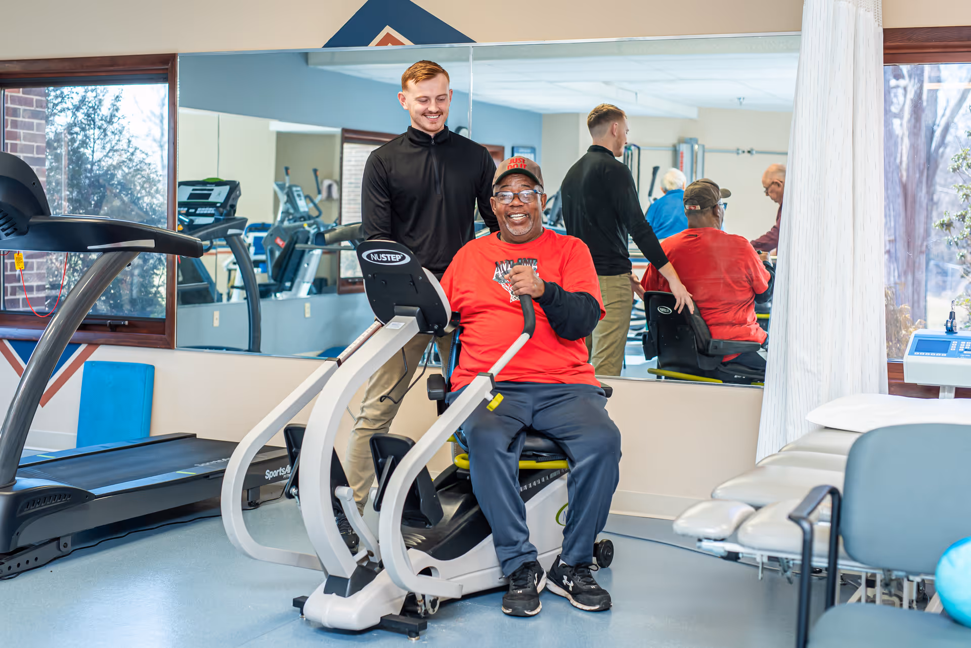 A senior man smiles on a recumbent exercise bike in a gym while a staff member assists him.