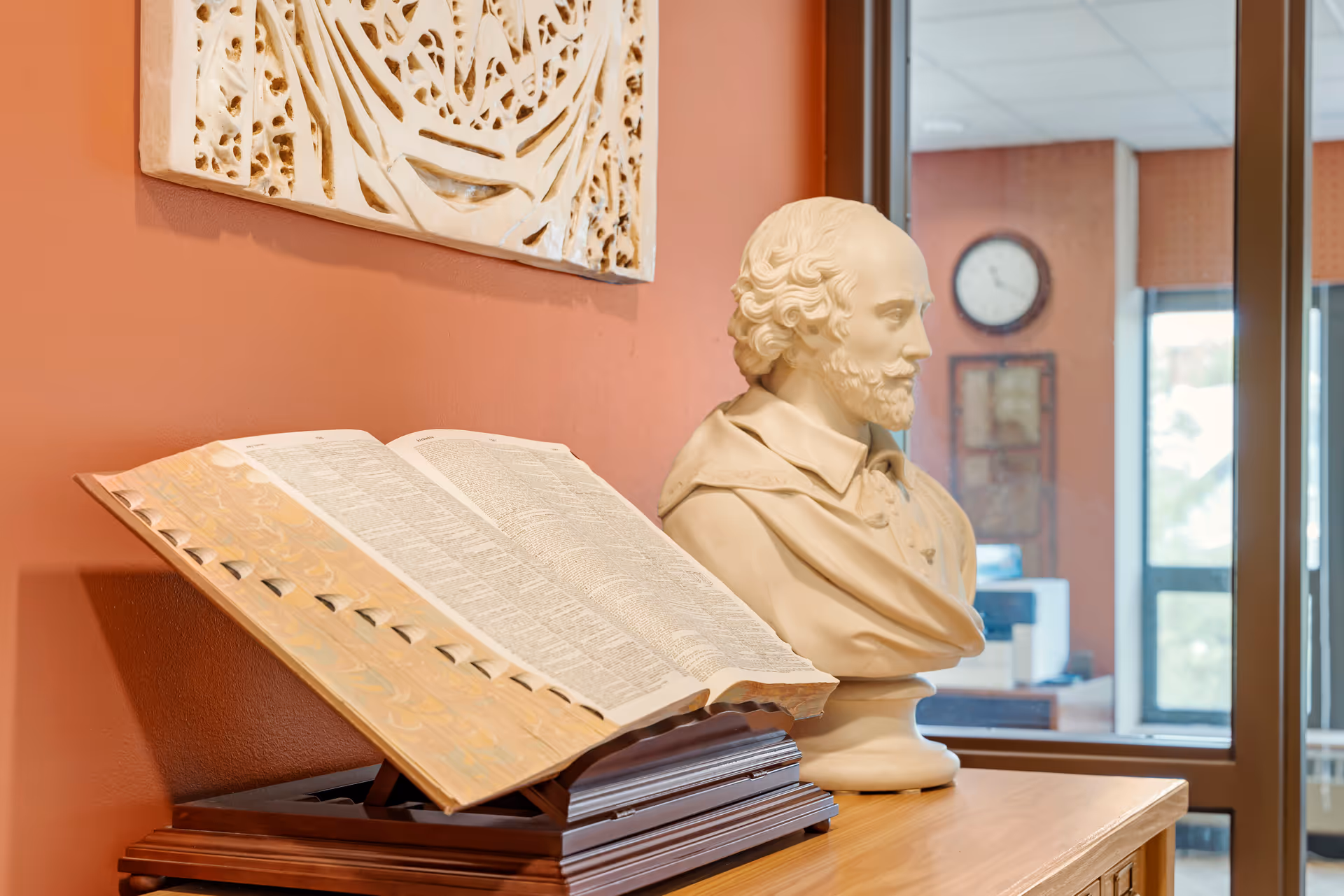 A bust sculpture of a bearded man with curly hair placed on a wooden surface next to an open large book on a stand, with a decorative wall hanging and a window showing a clock and part of an office in the background.