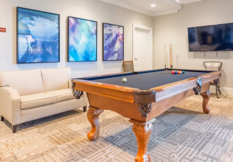 A recreation room with a wooden pool table with a black felt top, a beige loveseat, three abstract art paintings on the wall, a flat-screen TV mounted on the wall, and a chair with pool cues in the corner.