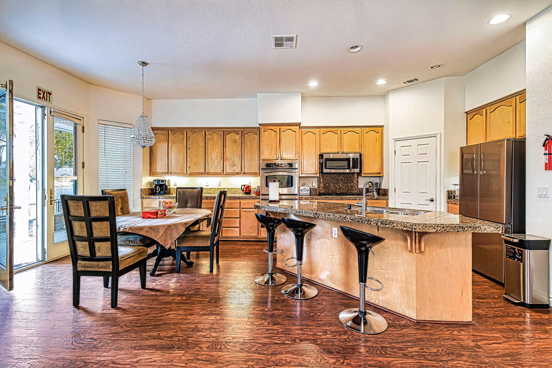 A spacious kitchen and dining area with wooden cabinets, granite countertops, and hardwood floors. The kitchen features a large island with three black bar stools, a built-in oven, microwave, and a stainless steel refrigerator. A dining table with six chairs is set near a glass door leading outside. The room is well-lit with recessed lighting and a hanging chandelier above the dining table.