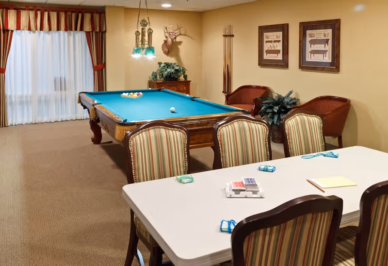 A game room with a pool table in the center, a set of pool cues mounted on the wall, and a mounted deer head above a small wooden cabinet with a plant. In the foreground, there is a table with striped upholstered chairs around it, playing cards and score pads on the table. The room has beige walls, carpeted floor, and a window with striped curtains.