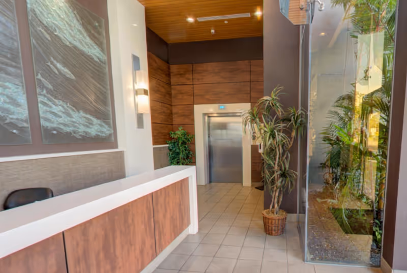Reception area with a wooden front desk, a chair behind it, large wall art depicting ocean waves, potted plants, an elevator with closed doors, and a glass wall showing greenery outside.