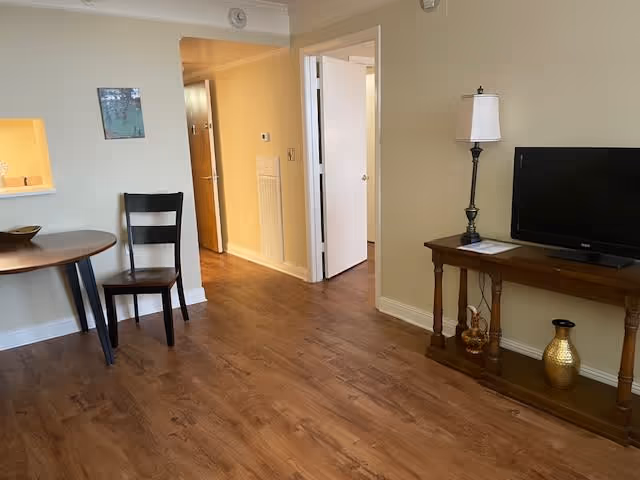 Interior view of a living area with wooden flooring, a small round table with a chair, a hallway leading to other rooms, and a wooden console table with a lamp, decorative items, and a flat-screen TV.
