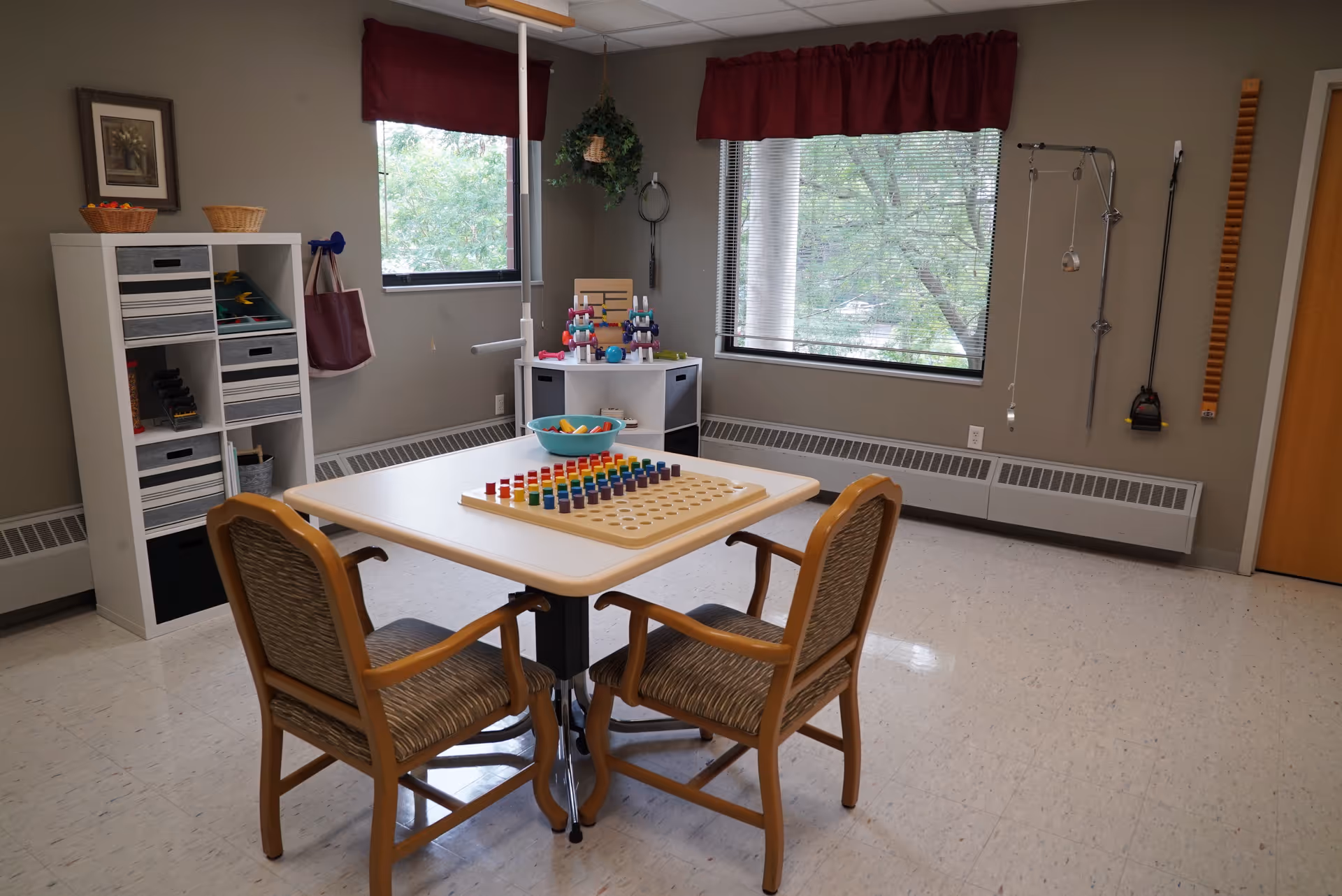 A well-lit activity room with two wooden chairs around a square table featuring a pegboard game with colorful pegs. The room has two windows with red valances, a white shelving unit with baskets and games, and various therapy or exercise equipment mounted on the walls. The floor is tiled, and the walls are painted a neutral color.