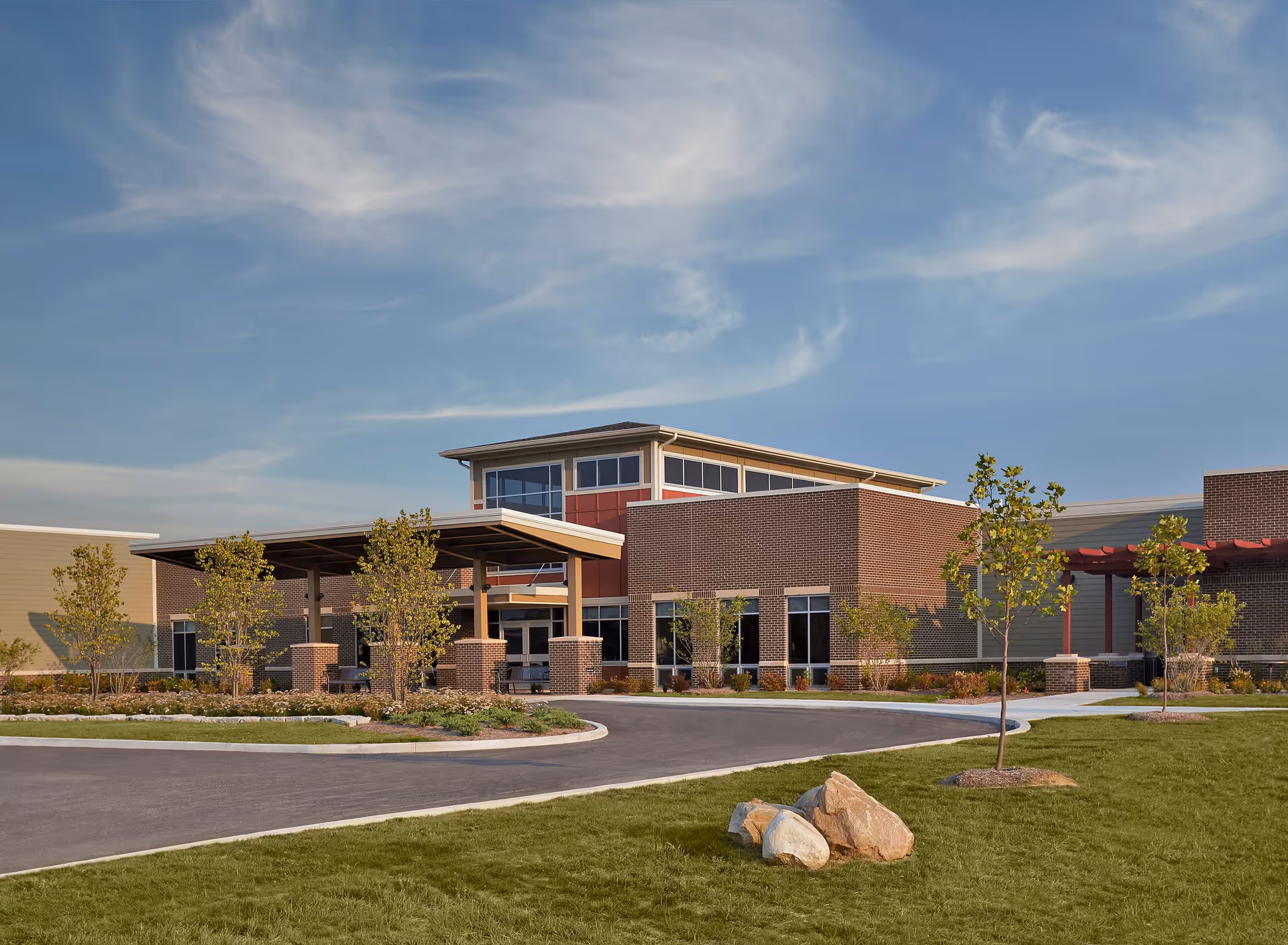 Front exterior of a modern brick senior living building with a covered entrance, driveway, young trees and a grassy lawn.