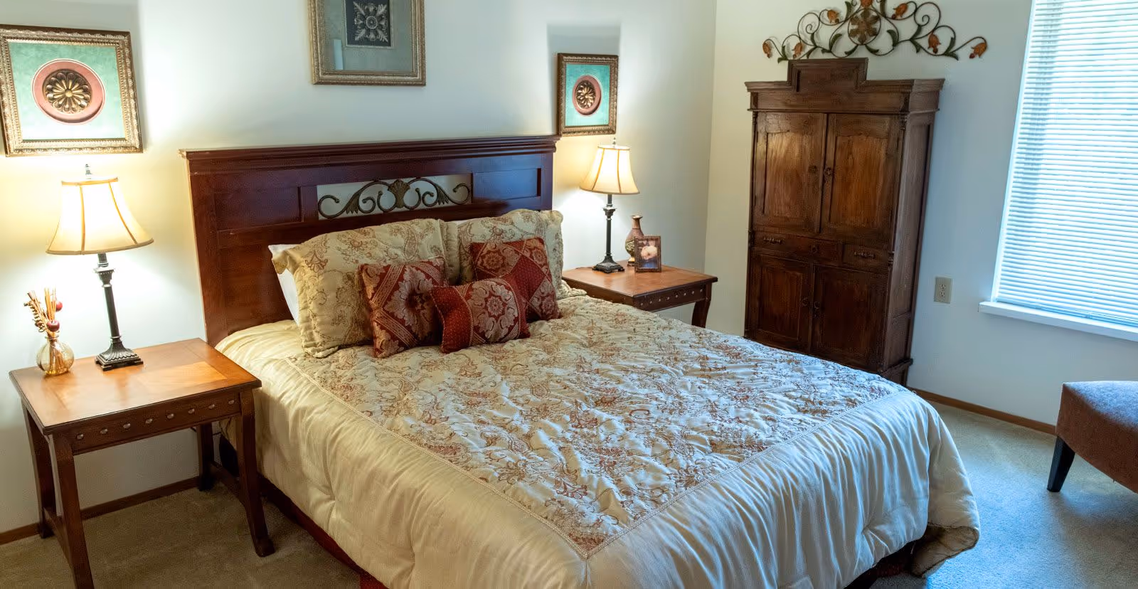 A cozy bedroom with a large bed featuring a decorative wooden headboard and patterned bedding with multiple pillows. There are two wooden nightstands on either side of the bed, each with a lamp. The room also includes a wooden armoire, a window with blinds, and framed artwork on the walls.