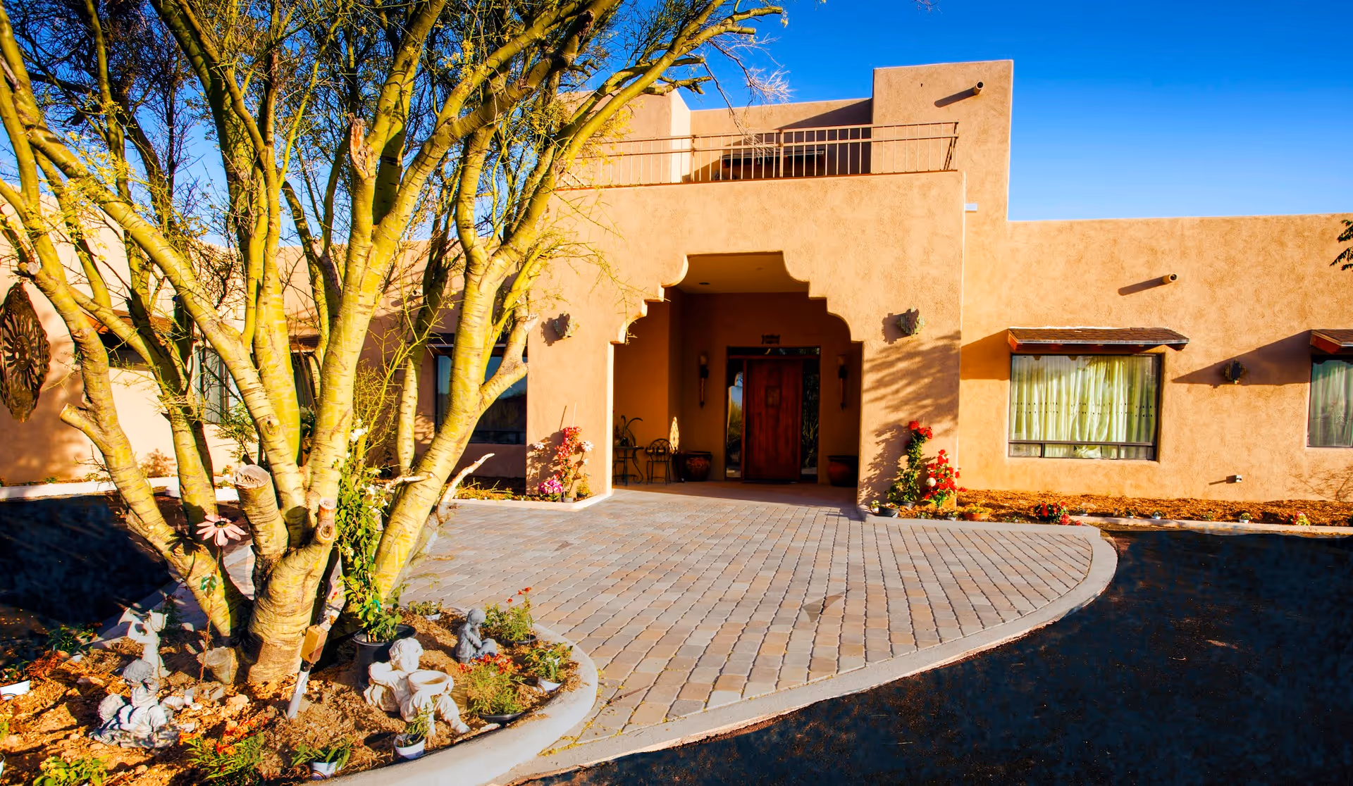Southwestern-style building entrance with a paved circular driveway, large tree and desert landscaping under a clear blue sky.